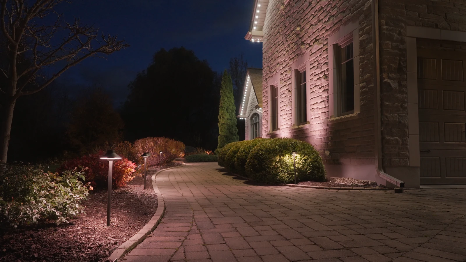 A curved stone walkway at night with path lights, lit shrubs and a soft glow on the stone wall of the home