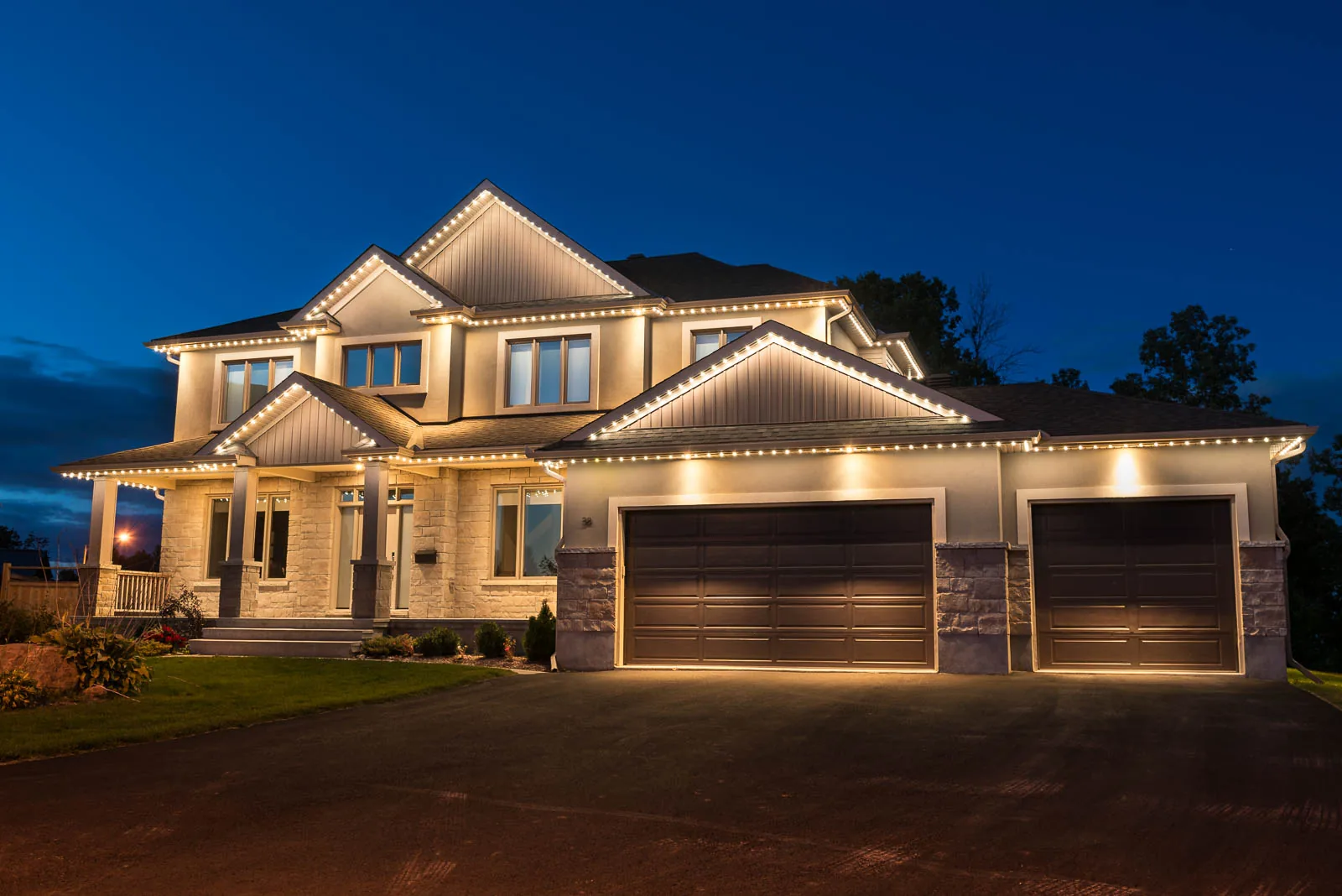 A two story home with warm white roofline lighting over a three car garage at dusk