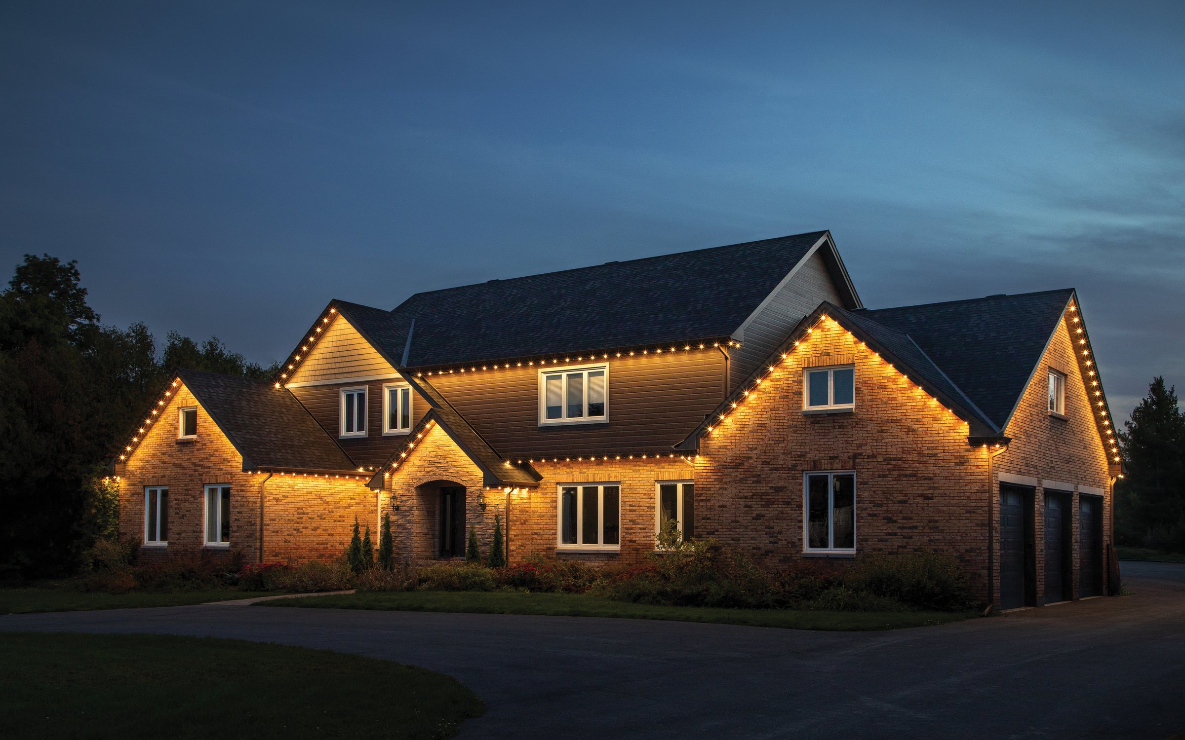 A large brick home at dusk with warm white permanent lighting tracing every gable and eave